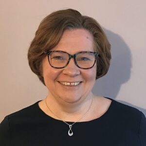 The head of a woman facing the camera and smiling. She has short hair, glasses and a black top