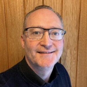 The head of a man facing the camera. He has short hair, glasses and wears a black shirt and sweater