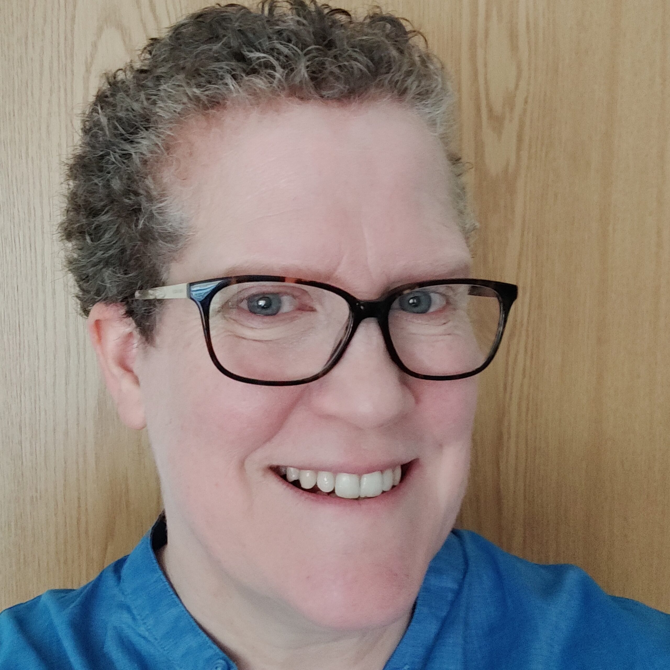 The head of a woman looking at the camera and smiling. She has short curly hair, glasses and a blue shirt