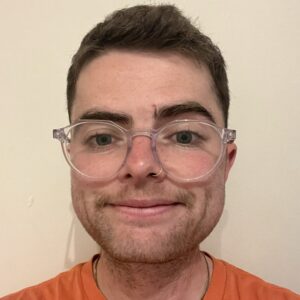 The head of a person facing the camera. He has short hair, wears glasses and is wearing an orange top.