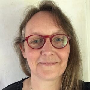 The head of a woman facing the camera. She has long hair and wears red glasses and a black top.