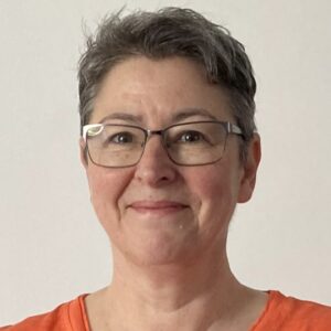 The head of a woman facing the camera and smiling. She has short hair, glasses and wears an orange top
