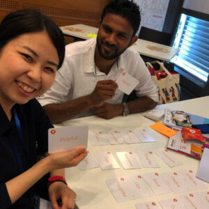 A man and a woman sit around a table and look at the camera. One holds a card saying 'playful'