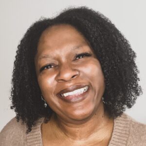 The head of a woman facing the camera and smiling. She has shoulder-length hair and wears a pale brown top