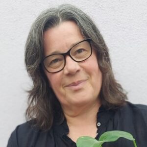 The head of a woman facing the camera and smiling. She has long hair, glasses and wears a black shirt. She's holding a plant.