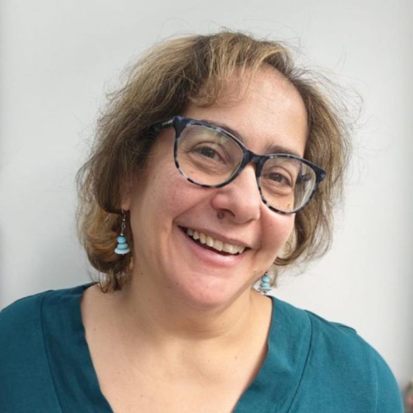 A woman smiling at the camera with her head titlted to one side. She has glasses, shoulder length hair and wears a teal green top.