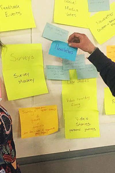 Two women standing in front of a board covered with large post-its during an Evaluating Community Development training session. They're both looking at the camera