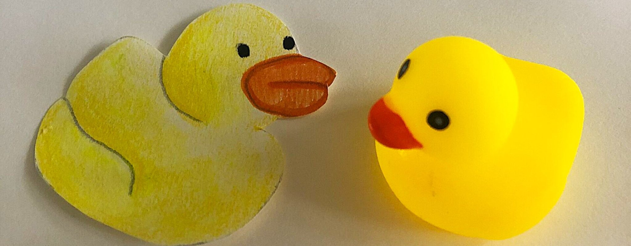 Yellow paper duck on a piece of paper with a yellow plastic duck next to it. They appear to be looking at each other.
